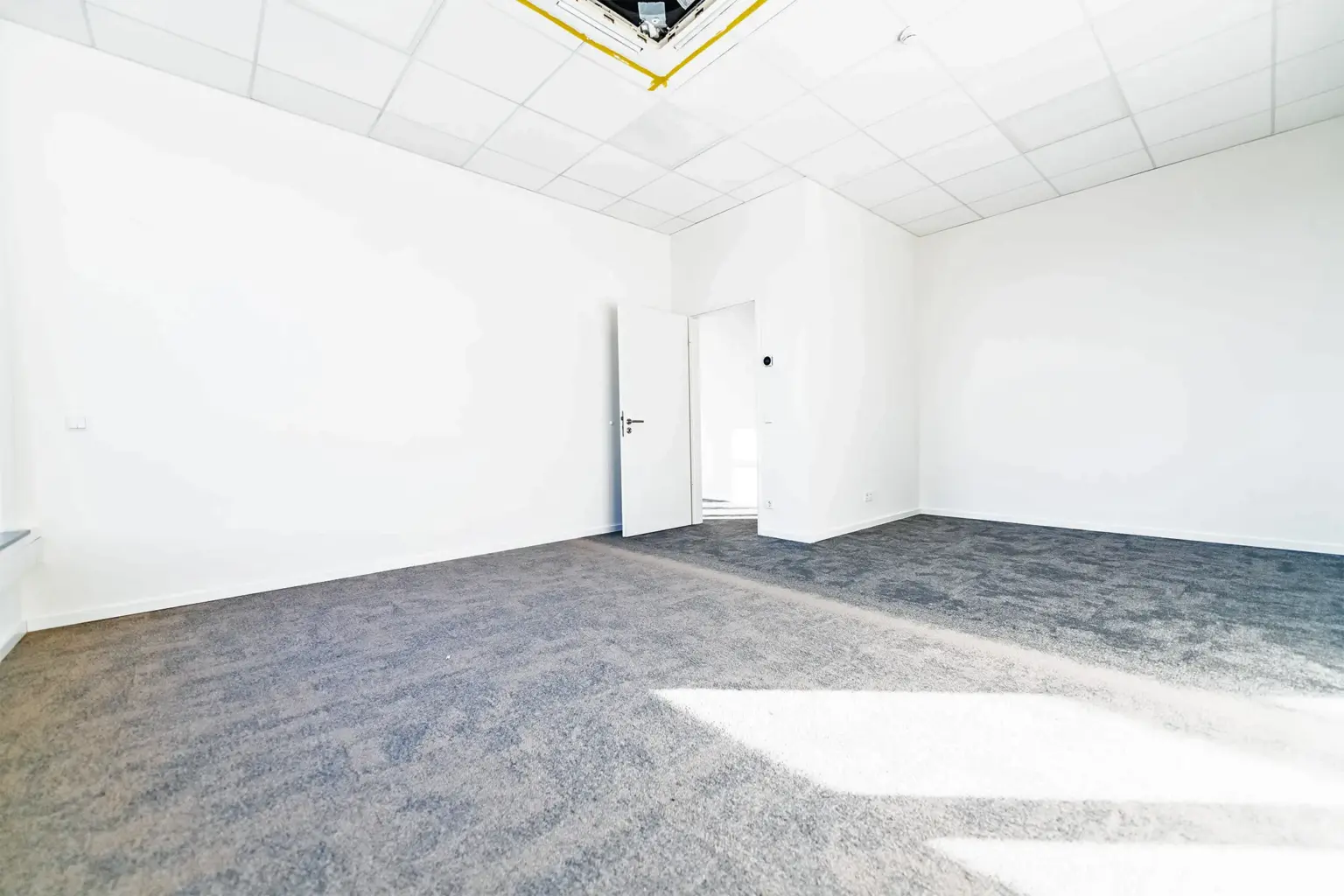 Leerer Büroraum mit weißen Wänden, grauem Teppichboden und Deckenleuchte.