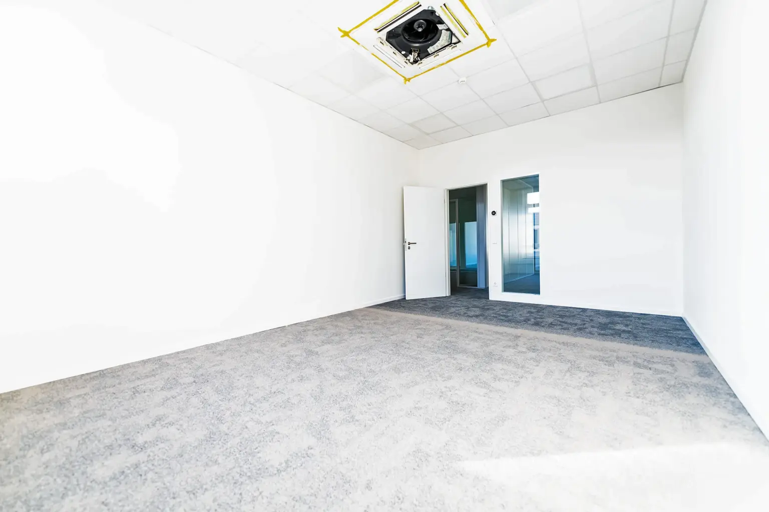 Leerer Büroraum mit weißen Wänden, Teppichboden und Deckenlicht.